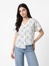A woman modeling a white short-sleeve button-up blouse with a delicate green and orange floral print, paired with light blue jeans, against a plain white studio background.