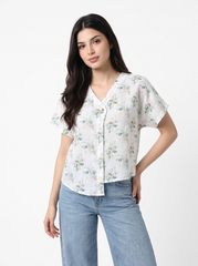 A woman modeling a white short-sleeve button-up blouse with a delicate green and orange floral print, paired with light blue jeans, against a plain white studio background.