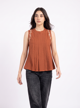A woman modeling a sleeveless rust-orange top with vertical pleats and light beige floral embroidery on the shoulders, paired with dark gray jeans, against a plain white studio background.