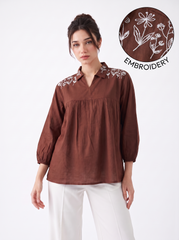 A woman models a brown long-sleeve blouse with white floral embroidery on the shoulders, paired with white pants. A circular inset in the top right shows a close-up of the brown fabric with white embroidery and the word 'EMBROIDERY'.