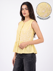 A woman wearing a sleeveless light yellow top with white floral embroidery on the front, paired with dark gray jeans. A circular inset in the top right shows a close-up of the embroidery detail with the text 'EMBROIDERY'.