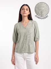 A woman wearing a sage green short-sleeve blouse with white floral embroidery on the front, paired with white pants. A circular inset in the top right shows a close-up of the embroidery detail with the text 'EMBROIDERY'.