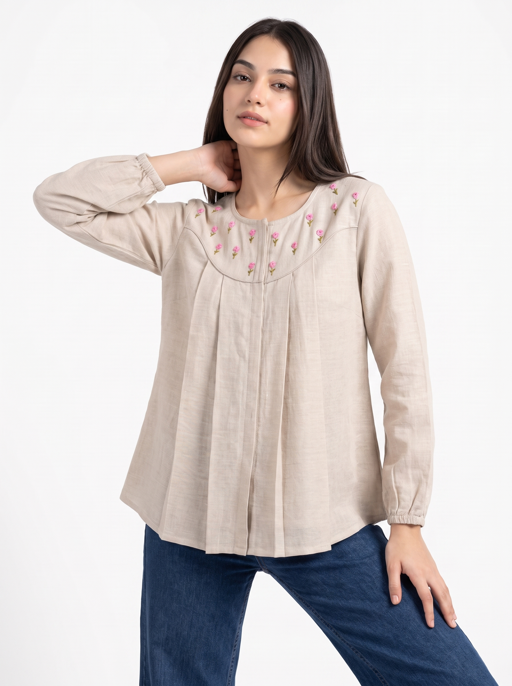 A woman modeling a light beige linen long-sleeve blouse with pink floral embroidery around the neckline, paired with blue jeans, against a plain white background.