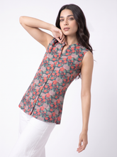 A woman modeling a sleeveless, mandarin-collar button-up top with a dense floral print in coral pink, red, and muted green on a white studio background.
