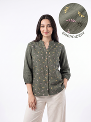 A woman modeling a sage green long-sleeve button-up shirt with a delicate multicolor embroidered pattern, paired with beige trousers. A circular inset in the top right highlights the embroidery detail.
