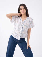A woman modeling a short-sleeve button-up blouse with a delicate blue and beige botanical print on a white base, paired with dark blue jeans, against a plain white studio background.