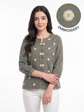 A woman wearing a sage green linen top with white and yellow daisy embroidery, paired with white pants. An inset circle in the top right shows a close-up of the embroidery detail with the text 'EMBROIDERY'.