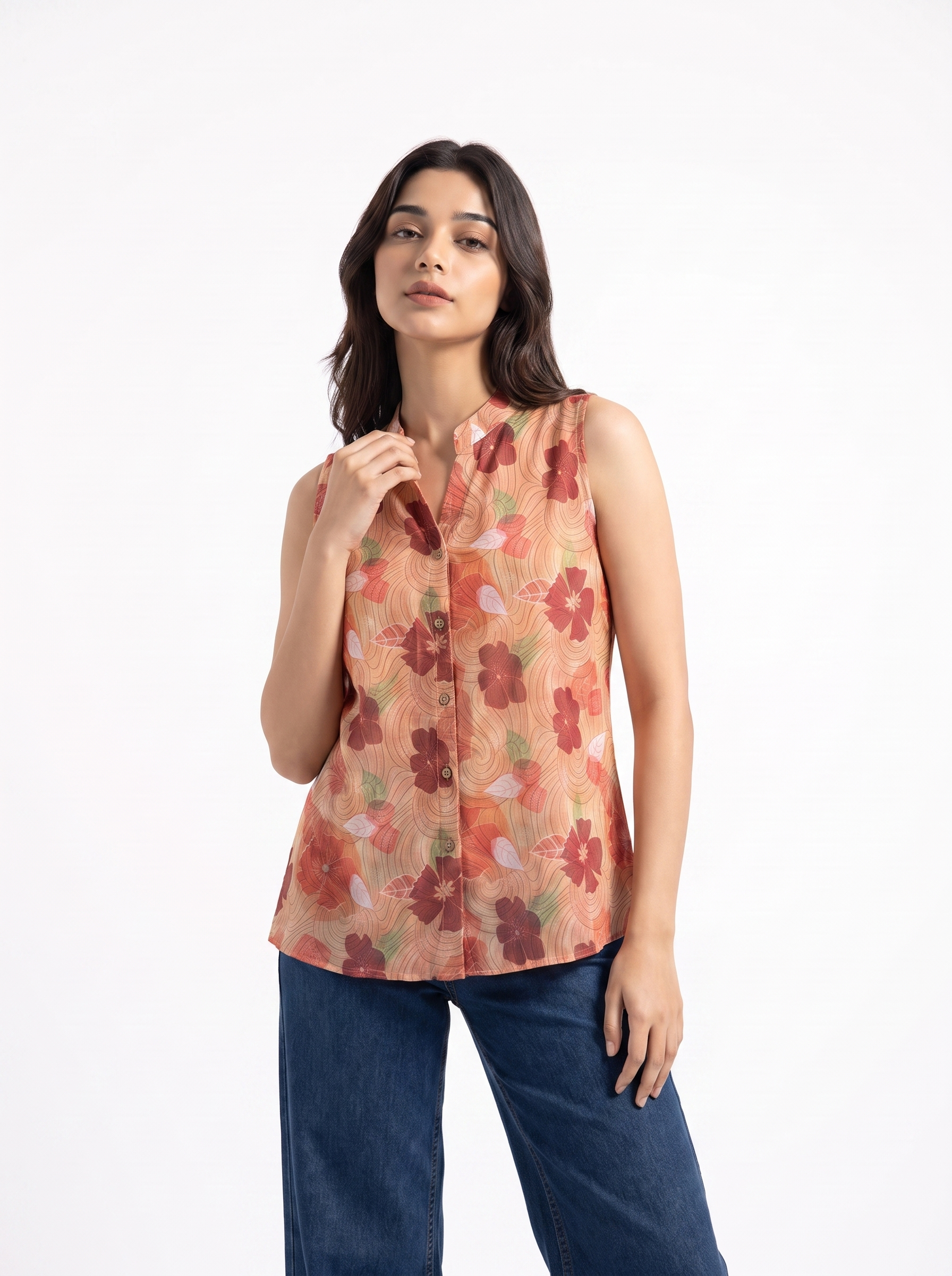 A woman modeling a sleeveless button-front blouse with a warm floral print in orange, red, and beige tones, paired with blue jeans against a clean white studio background.