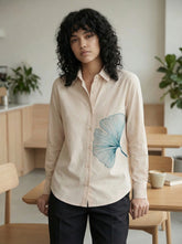 A refined cream shirt top featuring a striking teal ginkgo leaf graphic on the front for a clean artistic look.