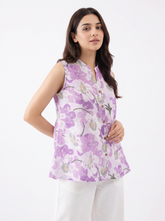 A woman modeling a sleeveless lavender floral printed top with a split neckline and button details, paired with white pants, against a plain white studio background.