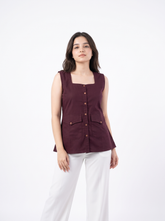 A woman modeling a sleeveless square-neck top in deep burgundy with brown buttons, paired with white pants, against a plain white studio background.