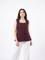 A woman modeling a sleeveless square-neck top in deep burgundy with brown buttons, paired with white pants, against a plain white studio background.