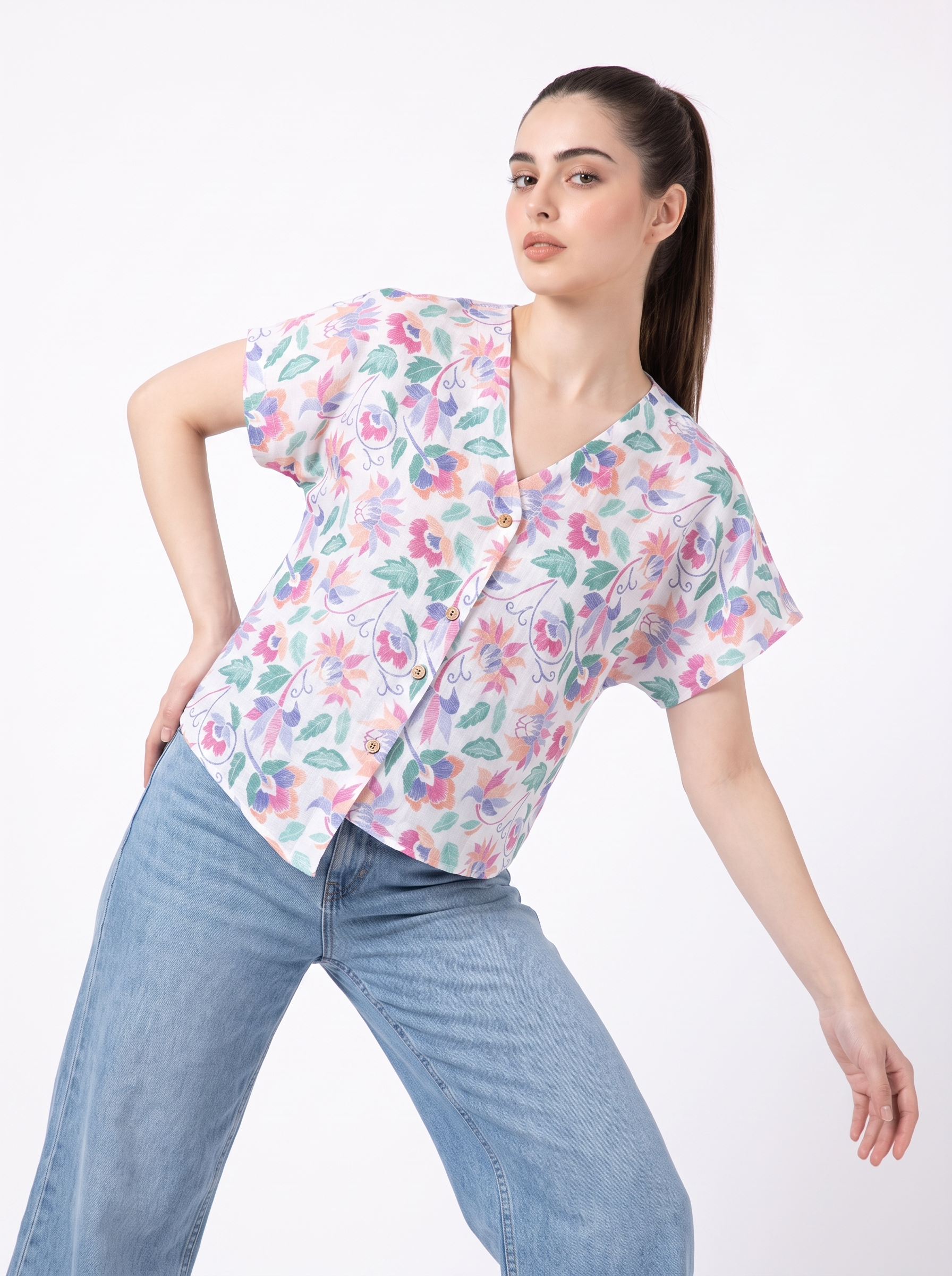 A woman modeling a short-sleeve V-neck button-up scrub top with a colorful floral print, paired with light blue jeans, against a plain white background.