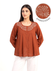 A woman wearing a rust-orange long-sleeve top with white embroidered detailing around the neckline, paired with white pants. An inset circle in the upper right highlights the embroidery texture with the word 'EMBROIDERY' on a white background.