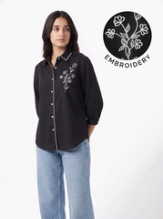 A woman modeling a black long-sleeve button-up shirt with white floral embroidery and white piping along the front placket, paired with light blue jeans. A circular graphic overlay in the top right shows a close-up of the white floral embroidery on black, labeled 'EMBROIDERY'.