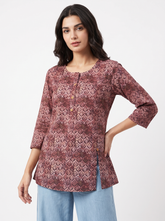 A woman modeling a three-quarter sleeve tunic top with a geometric pattern in dusty rose and burgundy, paired with light blue jeans, against a plain white studio background.
