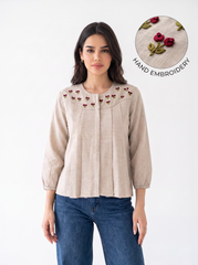 A woman wearing a beige linen blouse with hand-embroidered cherry motifs on the yoke, paired with blue jeans. An inset circle in the top right highlights the detailed cherry embroidery.
