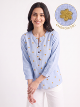 A woman wearing a light blue long-sleeve top with small gold embroidered dots, paired with white pants, against a plain white background. An inset circle in the top right shows a close-up of the gold embroidery texture with the word 'EMBROIDERY'.
