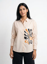 A woman modeling a light beige long-sleeve button-up shirt with a black and orange abstract floral graphic on the front, paired with black pants, against a plain white studio background.