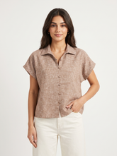 A woman modeling a short-sleeve button-front collared blouse in a light taupe-beige textured fabric, paired with white pants, against a plain light gray studio background.