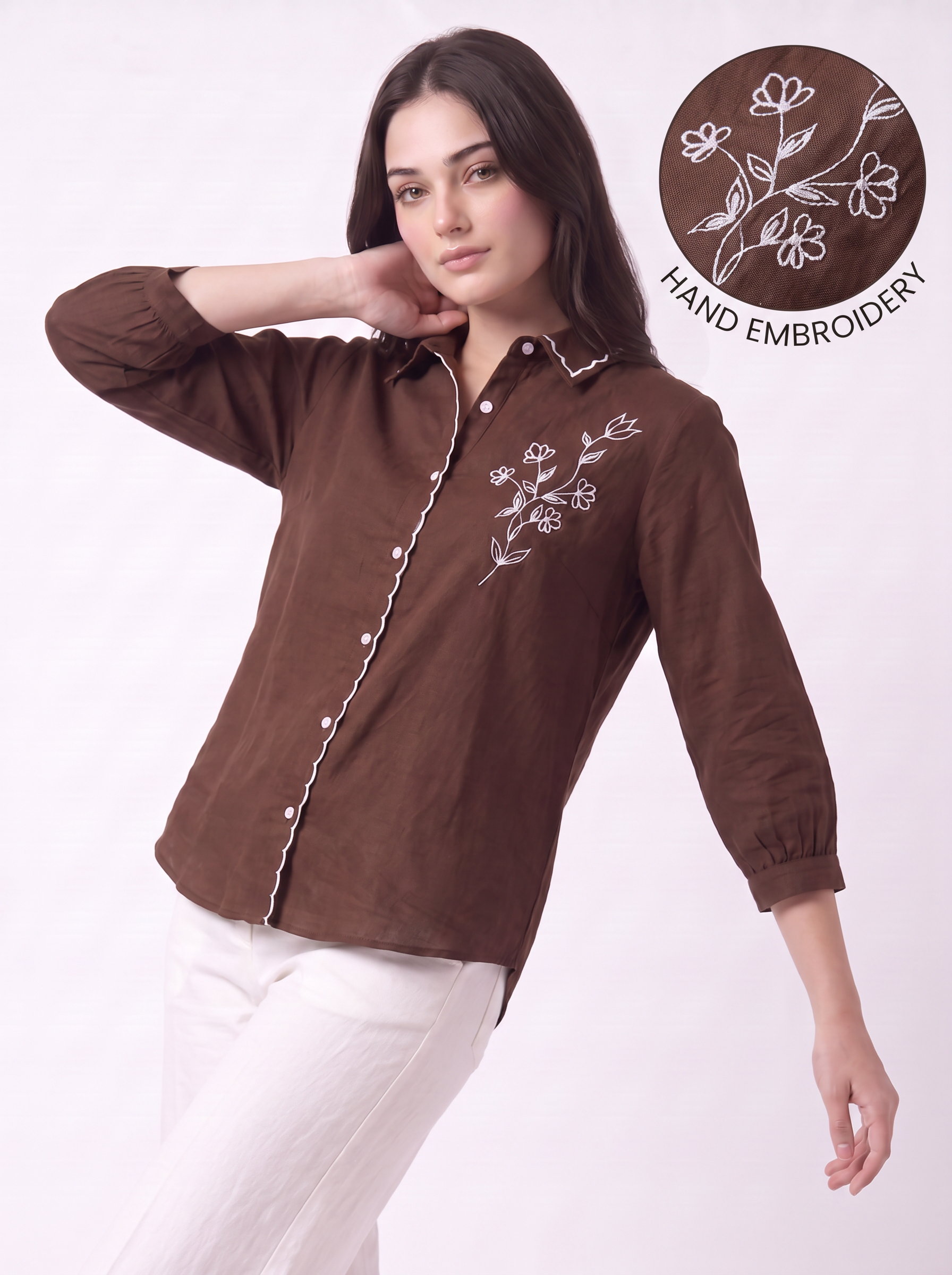 A woman modeling a brown long-sleeve button-up shirt with white floral hand embroidery on the chest and white piping along the placket and cuffs, paired with white pants. A circular inset in the top right highlights the hand embroidery.