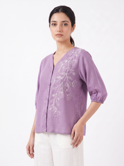 A woman modeling a lavender purple linen blouse with white floral embroidery on the front, paired with white trousers, against a plain white background.