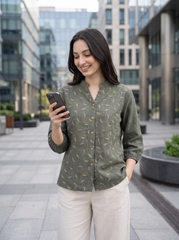 A woman standing outdoors in a modern city plaza, looking at her phone. She is wearing a sage green long-sleeve button-up shirt with a small yellow and white leaf pattern, paired with light beige trousers.