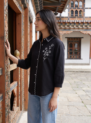 A woman wearing a black long-sleeve button-up shirt with white floral embroidery and white piping, paired with light blue jeans, standing in front of a traditional wooden building.