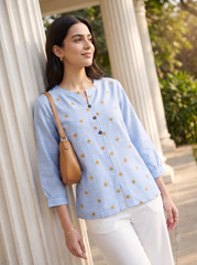 A woman wearing a light blue three-quarter sleeve kurta with small gold polka dots and white pants, leaning against a white column in an outdoor setting.