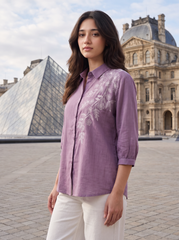 A woman wearing a lavender-purple linen button-up shirt with white floral embroidery on the front, standing outdoors in front of the Louvre Museum and glass pyramid.