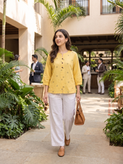 A woman walking in a tropical resort courtyard wearing a yellow embroidered kurta, white wide-leg trousers, and brown loafers, carrying a brown handbag.