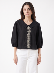 A woman modeling a black long-sleeve blouse with a Peter Pan collar and vertical floral embroidery down the front, paired with white pants, against a plain white studio background.