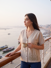 A woman stands on a balcony overlooking water and boats, wearing a light beige short-sleeve button-front cardigan with embroidered details and holding a small beige mug.
