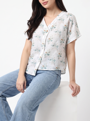 A woman wearing a short-sleeve button-front blouse with a delicate floral print in muted green, blue, and orange on an off-white base, paired with light-wash blue jeans, seated against a plain white background.