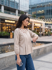 A woman wearing a beige linen long-sleeve blouse with red floral embroidery on the chest and blue jeans, standing in an outdoor urban plaza with a restaurant and glass building in the background.