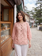 A woman stands on a cobblestone street wearing a light pink embroidered kurta with white pants. The kurta features small green and brown floral embroidery and a buttoned front.