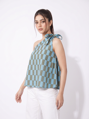 A woman modeling a sleeveless halter-style top with a blue and olive green checkerboard pattern and a tie detail at the shoulder, paired with white pants against a plain white studio background.