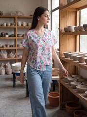 A woman wearing a short-sleeve floral V-neck blouse and light blue jeans walks through a pottery studio filled with shelves of ceramic bowls and pots.