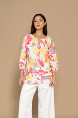 A woman modeling a three-quarter sleeve top with a colorful abstract floral print in yellow, red, and gray on a white base, paired with white wide-leg pants, set against a solid beige background.
