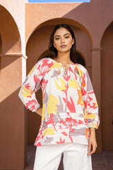 A woman modeling a long-sleeve tie-neck blouse with a colorful abstract floral pattern in red, yellow, and gray on a white base, paired with white pants, set against a warm terracotta archway background.