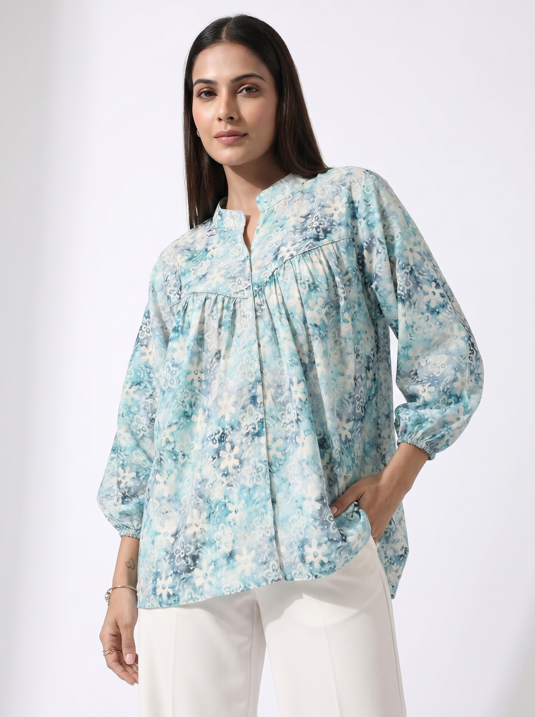A woman modeling a light blue and white floral print blouse with a split neckline and gathered sleeves, paired with white pants, against a plain light gray studio background.