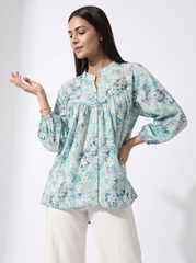 A woman modeling a light blue and white floral print long-sleeve blouse with a split neckline, paired with white pants, against a plain white studio background.