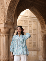 A woman wearing a light blue and white floral printed kurta with white trousers, standing in front of an ornate stone archway.