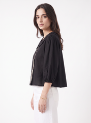 A woman wearing a black cropped puff-sleeve blouse with gold trim down the front, paired with white trousers, photographed against a plain white studio background.