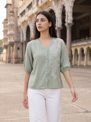 A woman wearing a sage green blouse with white floral embroidery on the front and white pants, standing in front of a historic stone building with arches.