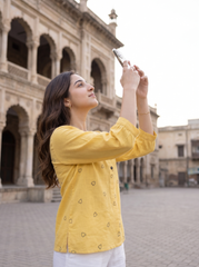 A woman in a yellow patterned linen shirt takes a photo of a historic stone building in a square.