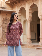 A woman wearing a dusty rose printed kurta and light blue jeans stands in front of a sandstone palace courtyard with arches.