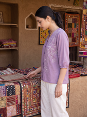 A woman in a lavender embroidered kurta and white trousers stands in a rustic shop, looking down at a table covered with colorful patchwork textiles.