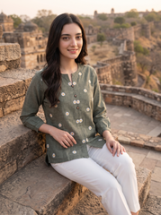 A woman sitting on stone steps at a historic fort, wearing an olive green linen kurta with white floral prints and white trousers.