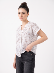 A woman modeling a short-sleeve button-up blouse with a delicate beige and brown botanical leaf pattern on a white base, paired with dark gray jeans, against a plain white studio background.